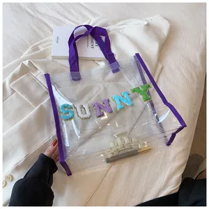 Detailansicht der transparenten PVC-Tasche mit bunten Buchstaben-Patches und violetten Henkeln.