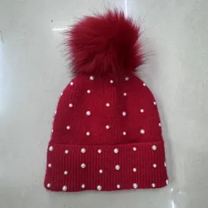 Detail shot of a winter warm red knit beanie hat featuring faux pearl studs and a fluffy pom pom.