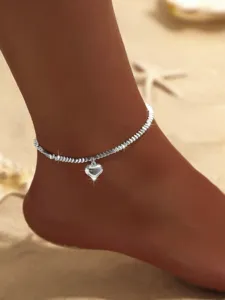 Nahaufnahme einer glitzernden silbernen Perlenfußkette mit Herzanhänger am Sandstrand.