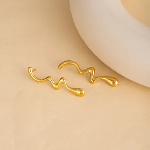 Detail shot of irregular wavy gold-colored stainless steel earrings on a textured beige surface.