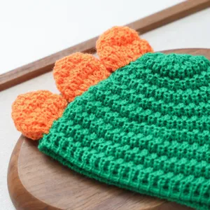 Gros plan détaillé du bonnet pour animal en crochet vert avec crêtes de dinosaure orange.