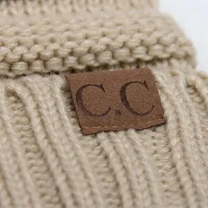 Close-up detail of beige knit beanie hat with brown leather brand tag, soft texture.