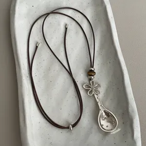 Brown cord necklace featuring a metallic spoon pendant, flower charm, and tiger's eye beads.