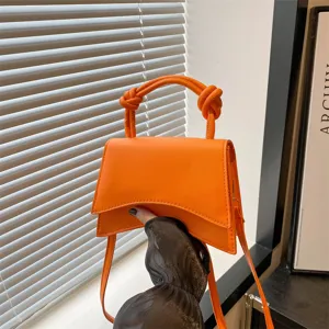 Stylish orange top handle crossbody bag with unique curved flap and chic knotted handles.