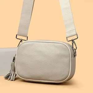 Beige leather camera crossbody bag featuring a wide woven strap and a stylish tassel charm.