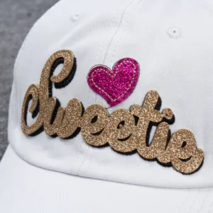 Close-up detail of white baseball cap with gold glitter Sweetie letter and pink heart embroidery.