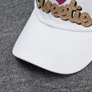 Detail shot of a white baseball cap featuring sparkling gold Sweetie embroidery and pink heart.