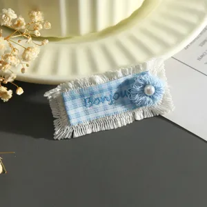 Dettaglio etichetta in tessuto scozzese blu e bianco, ricamo con frange e fiore di perla.