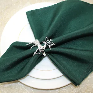 Detailed shot of a silver geometric reindeer napkin ring holding a dark green napkin on white plates.