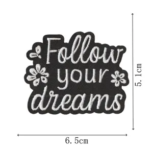 Detalle de parche bordado con tela negra, frase blanca motivacional y pequeñas flores.