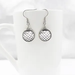 Detailed close up shot of silver fish scale pattern round dangle earrings displayed on white mug.