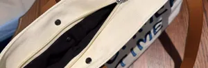 Canvas tote bag detail view showing zipper closure, blue embroidery and sturdy brown straps.
