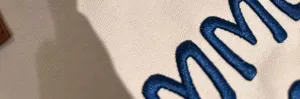 Canvas tote bag detail close-up showing blue 3D embroidery texture and premium fabric.