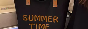 Black canvas tote bag detail, featuring embroidered yellow summer time letters and leather straps.