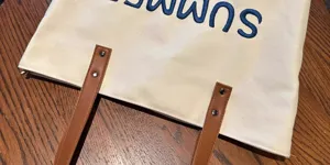 Canvas tote bag detail view, blue embroidered letters, brown PU handles, metal rivets.
