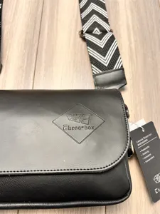 Detail view of black crossbody bag with wide geometric strap and branded logo stamp.
