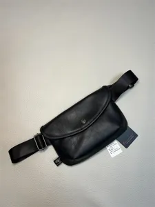 Detail view of black leather waist bag with metal snap, adjustable strap and branded tags.