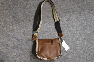 Close-up detail of casual brown leather messenger bag with striped webbing strap and zipper.