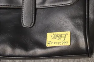 Close-up detail of black leather men business bag featuring brand logo patch, premium texture.