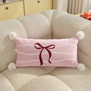 Pink lumbar pillow detail, soft velvet fabric with bow pattern and fluffy pom poms on sofa.