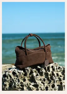 Detailed view of brown suede handbag with top handles and side pocket on rocky coastal scene.