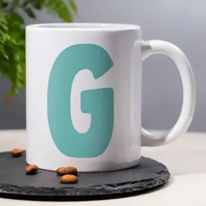 Close-up of a white ceramic coffee mug featuring a bold teal letter G design on slate.