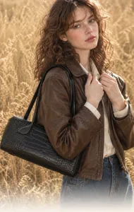 Detail view of black embossed leather shoulder bag styled with brown jacket in outdoor field.