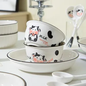 Set di stoviglie in ceramica con simpatici motivi di mucca, ciotola, piatto e cucchiaio.