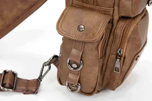 Close-up detail of brown leather sling bag with metal D-rings and zipper pockets.