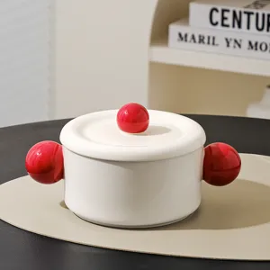 Ceramic soup bowl detail showing white body with red ball handles and lid knob on placemat.