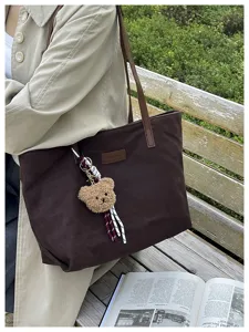 Detail view of the large brown canvas tote bag with cute plush teddy bear keychain worn outdoors.