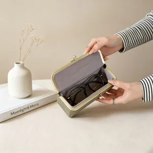 Detailed shot of olive green kiss-lock glasses case with soft grey interior lining on beige table.
