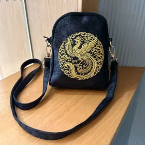 Detail shot of a black cross-body bag featuring exquisite gold phoenix embroidery and hardware.