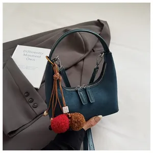 Detail shot of the dark blue suede dumpling bag showing its zipper, texture, and pom-pom charm.