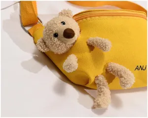 Close-up detail of the yellow canvas chest bag featuring plush bear decoration and zipper.