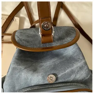 Close-up detail of casual denim backpack, brown leather trim, metal snap button, strap.