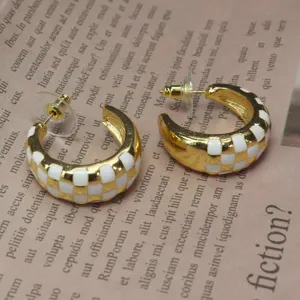 Gros plan de boucles d'oreilles créoles C émail à carreaux or et blanc sur journal.
