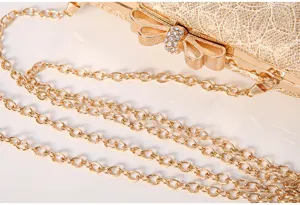 Gold lace evening bag detail showing rhinestone bow clasp and metal chain strap texture.