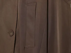 Brown trench coat fabric detail close-up showing button and pocket stitch texture.