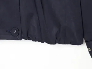 Close-up detail of dark navy blue short coat hem, featuring pleated design, button and metal eyelet.