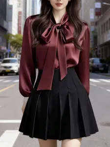 Detailed view of the burgundy satin ribbon bow blouse and black pleated skirt on street.