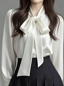 Close-up detail of elegant white satin bow tie blouse with long sleeves and pleated skirt.