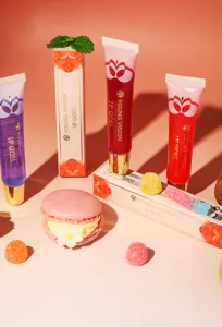Detail view of glossy lip gloss tubes and colorful boxes styled with sweet macarons and gummy candies.