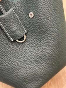 Close-up detail of dark green pebbled leather bucket bag with silver hardware and white stitches.