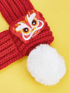 Close-up detail of red knitted pet scarf with white pom-pom and festive lion dance embroidery.