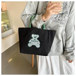 Black canvas tote bag detail with green striped bear patch and green handles for daily use.