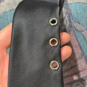 Close-up detail of black punk style necktie featuring three metallic eyelets and textured fabric.