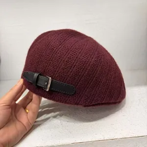 Close-up view of a wine red knitted beret hat featuring a stylish black leather strap buckle.