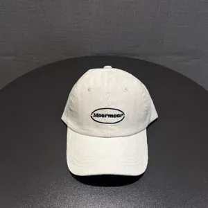 White corduroy baseball cap close-up featuring black embroidered oval logo lettering detail.