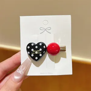 Close-up detail of black polka dot heart and red ball hair clip on white display card.